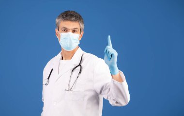 Doktor gibi giyinmiş olgun bir beyaz adam mavi arka planda duruyor. Yukarı bakıyor, maske ve eldiven takıyor, sağlık danışmanlığı yapıyor. İfadesi ciddidir ama yine de yakındır..