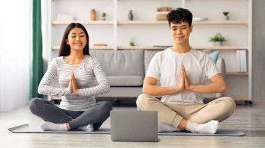 Evde sabah meditasyonu yapan rahat Asyalı bir aile, dizüstü bilgisayarın önünde Lotus 'ta poz veriyorlar, online sınıfları, oturma odaları var. Sportif çift birlikte egzersiz yapıyor.