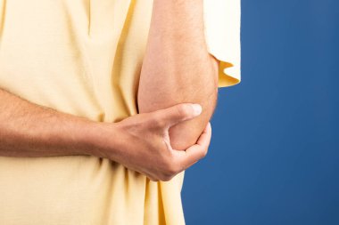 50 'li yaşlarda olgun bir beyaz adam yalnız başına oturuyor, dirseğini tutarken rahatsız görünüyor. Arka plan rahatlatıcı bir maviliktir. Yalnızlık ve yansıma anlarını çağrıştırır..