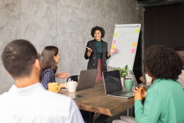 Uluslararası bir ofis ortamında, çeşitli bir ekip ortak bir toplantıya katılıyor. Bir kadın fikir sunarken, meslektaşlar anlayışı paylaşıyor, takım çalışmasını teşvik ediyor ve işbirliği yapıyor..