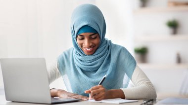 Uzaktan Öğrenme Konsepti. Sırıtkan İslami Kadın Başörtüsü Çalışmasında Evde Dizüstü bilgisayarla, Afrikalı Müslüman Kadın bilgisayardan ders alırken, uzaktan eğitimin keyfini çıkarıyor