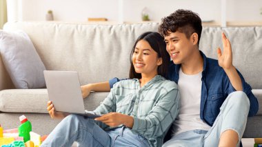 Oturma odasında yerde oturan, video görüşmesi yapan, bilgisayar kullanan, internet üzerinden ev temizleme hizmeti arayan neşeli Asyalı çift. Defterli yorgun genç adam ve kadın.