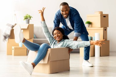 Neşeli evli siyah çift yeni evine taşınırken eğleniyor. Afro-Amerikan kadın oturma odasındaki karton kutuya biniyor. Neşeli eşler taşınmayı kutlarken eğleniyor.