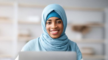 Siyah İslami Bağımsız Kadın Başörtüsü Altında Laptop kullanan ve kameraya gülümseyen, Müslüman Afrikalı Kadın İç İşleri Bakanlığı 'nda uzaktan bilgisayarla çalışıyor, Mesafe İşi' nin keyfini çıkarıyor,