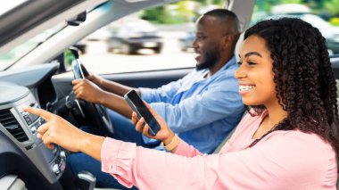 Yol Yolculuğu Konsepti. Profil yan görüntüsü: gülümseyen siyah bir çift lüks bir arabada oturuyor, erkek araba kullanıyor, cep telefonu olan kadın yola bakıyor, yol gösteriyor, dijital harita uygulaması kullanıyor.