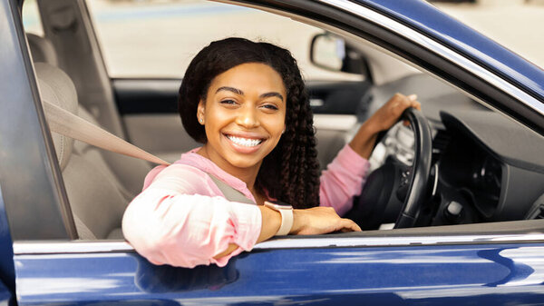 Позитивная молодая афроамериканка, сидящая за рулем. Счастливая черная женщина позирует перед камерой через открытую дверь автомобиля и улыбается. Автотест-драйв, вымывание, покупка, концепция проката