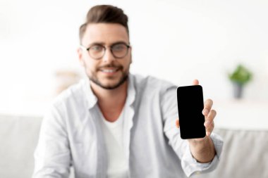 Bu web sitesini tavsiye ederim. Genç adam kameraya boş ekranlı akıllı telefonu gösteriyor. Reklamınız ya da tasarımınız için model alıyor. Evdeki koltukta oturan adam, elinde cep telefonu, seçici bir odaklanma.