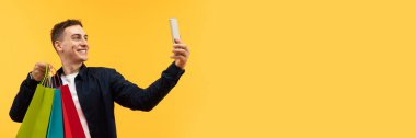 Şık genç adam telefonda selfie çekiyor, renkli kağıt poşetler tutuyor, sosyal medyada fotoğraf paylaşıyor, satış sezonunun keyfini çıkarıyor, sarı stüdyo arka planı, kopyalama alanı.