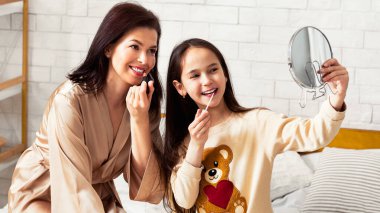 Evdeki güzellik salonu. Neşeli anne ve kızı yatağa aynalı ruj sürüyorlar.