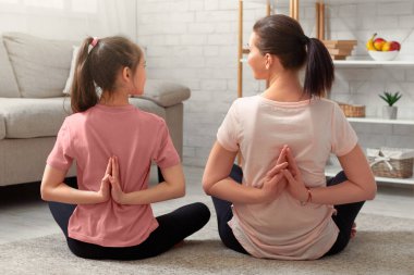 Genç anne ve kızının evde yoga yaparken, nilüfer çiçeği pozunda otururken, namaste jestleri yaparken. Ebeveynler ve çocuklar koronavirüs karantinası sırasında aile içi spor yapıyorlar.