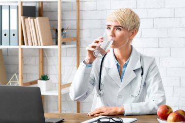 Doktor diyetisyen, iç kısımdaki ofiste laptoplu ve kırmızı elmalı camdan su içiyor.