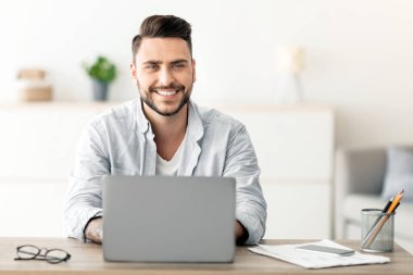 Başarılı bir serbest meslek sahibi. Mutlu adam iş yerinde dizüstü bilgisayarla oturuyor, gülümsüyor ve kameraya bakıyor, ev ofisindeki bilgisayarda uzaktan çalışıyor, boşluğu kopyalıyor. İnternet üzerinden uzak işten hoşlanan adam