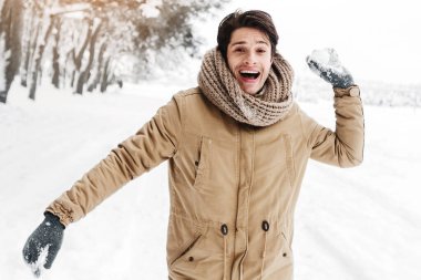 Kış eğlencesi. Kartopu atan mutlu genç adam kameraya bakıyor Ormanda karlı havanın tadını çıkarıyor