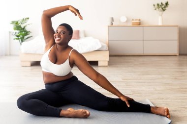 Kilolu siyahi bir kadın vücudunu esnetiyor yoga minderinin üzerine eğiliyor ve evdeki tecrit sırasında aktif bir yaşam sürüyor. Kıvrımlı genç kadın içeride çalışıyor.