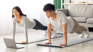 Evde dizüstü bilgisayar kullanan pozitif Asyalı bir çift var. Neşeli Çinli erkek ve kadın birlikte spor paspası çekiyorlar, bilgisayar ekranına bakıyorlar, video izliyorlar, panorama