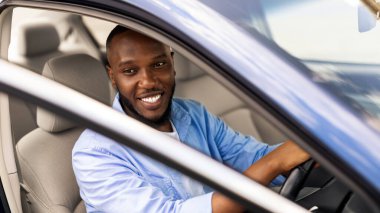 Pozitif Afro-Amerikalı genç bir adam araba kullanıyor ve açık otomobil kapısından bakıyor, park ediyor ya da yolcuyu arıyor. Otomatik test sürüşü, kiralama, kiralama, kiralama konsepti