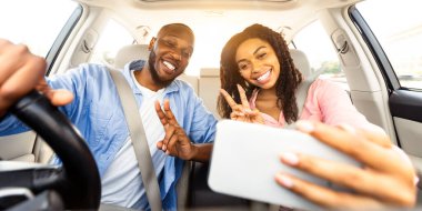 Güzel Afro-Amerikan çift arabada selfie çekiyorlar, cep telefonu kullanıyorlar, zafer işareti işareti gösteriyorlar. Mutlu siyah adam ve kadın tatile çıkıyor, otomatikman fotoğraf çekiyorlar.
