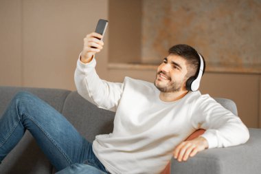 Gözlüklü genç bir beyaz adam evdeki bir kanepede rahatça oturup akıllı telefonuyla müzik dinlerken gülümsüyor. Doğal ışık sıcak atmosferi güçlendirir..