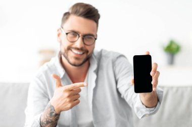 Akıllı telefon maketi. Milenyum adamı cep telefonunu boş ekranla gösteriyor ve parmağıyla işaret ediyor. İlan ya da tasarımın için yer var. Evdeki koltukta oturan adam, elinde telefonla.