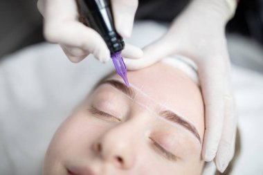 Şık bir salonda, bir kadın teknisyen olarak rahatlar ve kaşlarına kalıcı makyaj uygular. Atmosfer sakin, güzellik ve kişisel bakım prosedürlerine odaklanmış durumda..