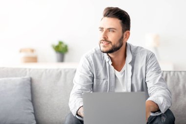 Milenyum serbest çalışanı laptopta çalışıyor ve bir kenara bakıyor, başarılı iş stratejisini düşünüyor ya da tatilin hayalini kuruyor, evdeki iç kısımda kanepede oturuyor, boş alan arıyor.
