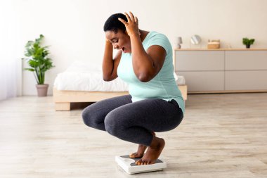 Artı boyut siyah kadın kilo verme sonuçlarına inanamıyor, bağırıyor, evde oturup tartıda oturuyor, başarıyı zayıflatmak için heyecanlanıyor, başarıyı kutluyor, kopyalama alanı yaratıyor. Panorama