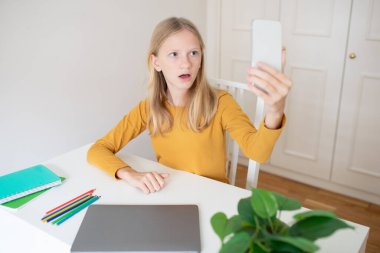 Genç bir kız rahat bir ev ortamında masasında oturuyor, akıllı telefonuyla video sohbeti yaparken şaşırıyor. Renkli kırtasiye malzemeleri ve dizüstü bilgisayar yakınlarda, canlı bir atmosfer yaratıyor..