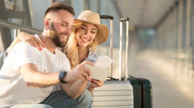 Tatil başlıyor. Mutlu çift biletlerle selfie çekiyor, havaalanı binasında uçak bekliyor.