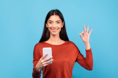 Uzun kumral saçlı bin yıllık bir bayan akıllı telefonuyla konuşurken mutlu bir şekilde gülümsüyor. Parlak mavi bir arka plana karşı poz veriyor, yaşam tarzında bir neşe ve bağlılık hissi sergiliyor..