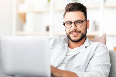 Evde dizüstü bilgisayar kullanan, oturma odasında kanepede oturan, gözlüklü yakışıklı bir adam. Evden çalışan, e-posta ya da programlama yazan mutlu bir adam.
