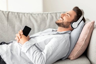 Kablosuz kulaklıklı mutlu adam kanepeye uzanıyor, gözleri kapalı ve gülümsüyor, evde müzik dinlerken, akıllı telefon tutuyor, yeni stereo kulaklıkların keyfini çıkarıyor.