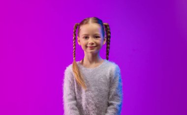 Örgülü örgülü örgülü neşeli bir kız canlı, mor bir arka plana karşı güven içinde oturur. Yumuşak bir kazak giyiyor ve modaya uygun, fütüristik bir hava yayıyor..