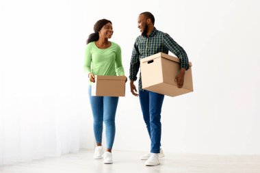 Karton kutular taşıyan mutlu Afrikalı Amerikalı çiftin portresi, mutlu siyah milenyum eşleri yeni dairelerinde pencerelerinin yanında yürüyorlar, birlikte hareket ediyorlar, boşluğu kopyalıyorlar.