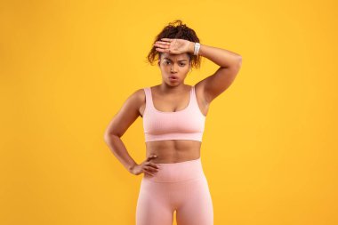 Antrenman kıyafeti giyen kararlı bir kadın bir eli kalçasında, diğer eli de alnında duruyor. İfadesi fitness ile ilgili bir aktivitede odaklandığını ve çaba gösterdiğini gösteriyor..