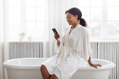 Parlak bir banyoda, küvetin kenarında oturan bir kadın, yumuşak bir bornoz giyiyor. Telefonuna bakarken gülümsüyor, evde bir anlık özen ve rahatlama anı kucaklıyor..