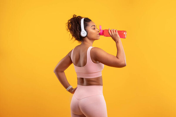 A woman dressed in workout gear drinks from a pink water bottle while listening to music through her headphones. The bright yellow background enhances her focus and energy during the workout session.