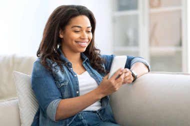 Genç siyah bir kadın rahat bir kanepede akıllı telefonunu kullanırken gülümsüyor. Parlak oturma odasında rahat bir dekor ve hafif ışıklandırma var. Rahat bir atmosfer yaratıyor..