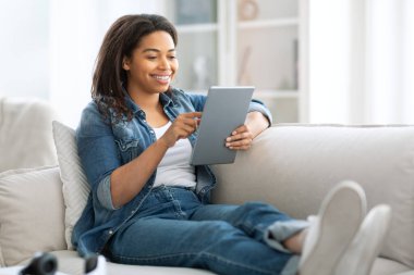 Genç siyah bir kadın rahat bir kanepede dinleniyor, tabletine bakarken gülümsüyor. Sıradan bir kot pantolon giyer ve yumuşak mobilyalarla süslenmiş iyi ışıklandırılmış bir oturma odasında vakit geçirmekten hoşlanır..