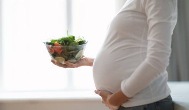 Hamile bir kadın parlak bir mutfakta karnını nazikçe kucaklarken elinde sebze ve sebzelerle dolu bir kâse tutarak hamilelik süresince sağlıklı beslendiğini belirtiyor.