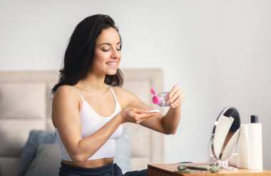 A young brunette woman is happily applying skincare products in her modern bathroom. The space is bright and inviting, enhancing her pampering experience. clipart