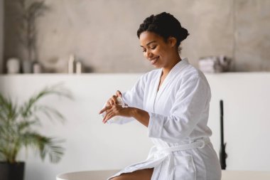 Beyaz bornozlu Afro-Amerikalı kadın spa 'da vakit geçirmekten zevk alıyor, gülümseyerek el kremi sürüyor. Sakin atmosfer ve doğal elementler onun rahatlama deneyimini arttırır..
