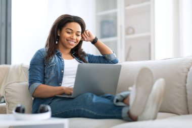 Genç bir kadın rahat bir şekilde hafif bir kanepede oturuyor, dizüstü bilgisayarıyla meşgul. Parlak gün ışığı odayı aydınlatıyor, rahat bir atmosfer yaratıyor..