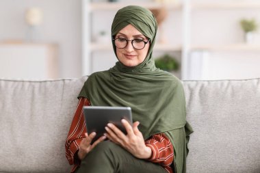 Yaşlı Müslüman kadın dijital tablet kullanıyor. İnternette kitap okuyor. Evdeki koltukta oturuyor. Modern Orta Doğu Ev Kadınları Başörtülü ve Gözlüklü Film İzliyor, İnternet Bilgisayarına Taranıyor