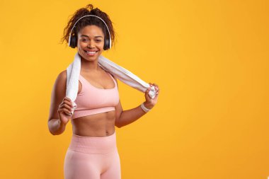 Bir kadın boynuna havlu tutarken gülümsüyor, spor kıyafetleri ve kulaklıklar takıyor. Parlak sarı arka plan enerjik bir atmosfer yaratır, boşluğu kopyala
