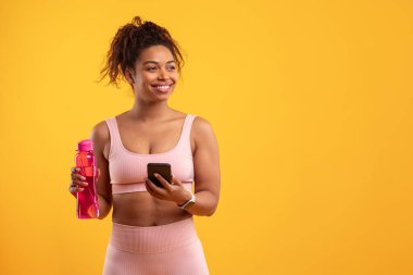 Pembe spor kıyafetleri giymiş bir kadın, parlak sarı bir zemine karşı duruyor. Bir elinde akıllı bir telefon ve pembe bir su şişesi tutuyor, pozitif ve enerjik bir titreşim yayıyor, uzayı kopyalıyor.