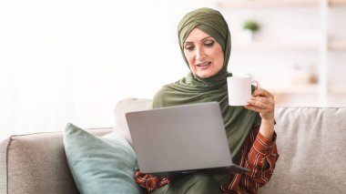 Modern Müslüman kadın dizüstü bilgisayar kullanıyor, sabah kahvesini içiyor, internetten film izliyor ve evdeki koltukta oturmuş haberleri okuyor. Bilgisayar aracılığıyla kıdemli İslami Ev Kadınları Ağı. İnsanlar ve Gadget