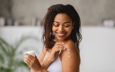 Bir kadın, kapalı bir alanda omzuna krem sürerken gülümser. Bir elinde küçük bir kap tutuyor ve cilt bakımının keyfini çıkarıyor..