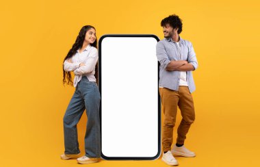 Mobile offer. Millennial indian couple leaning on giant smartphone with mockup, advertising or promoting app or website over yellow studio background