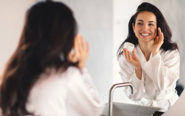 Genç esmer bir kadın banyoda cilt kremi sürmekten mutlu. Yansımasına gülümsüyor. Rahatlatıcı bir ev ortamında güzelliği ve kendine bakımı somutlaştırıyor..