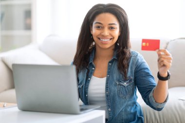 Genç siyah bir kadın parlak oturma odasında bir masada oturuyor, elinde bir kredi kartıyla mutlu bir şekilde dizüstü bilgisayarına bakıyor. Rahat bir ortamda online alışverişin keyfini çıkarıyor..